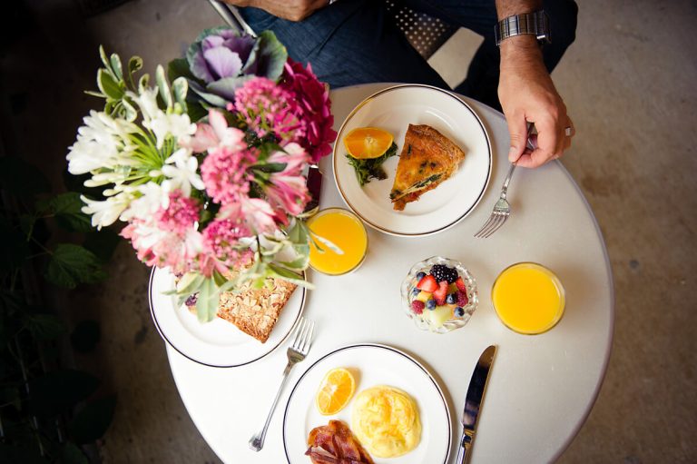 A table with breakfast plates, orange juice, a fruit bowl, and a bouquet of pink and white flowers.